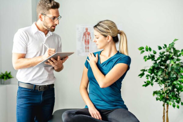 Male chiropractor listening to a female patient pointing to her sore shoulder