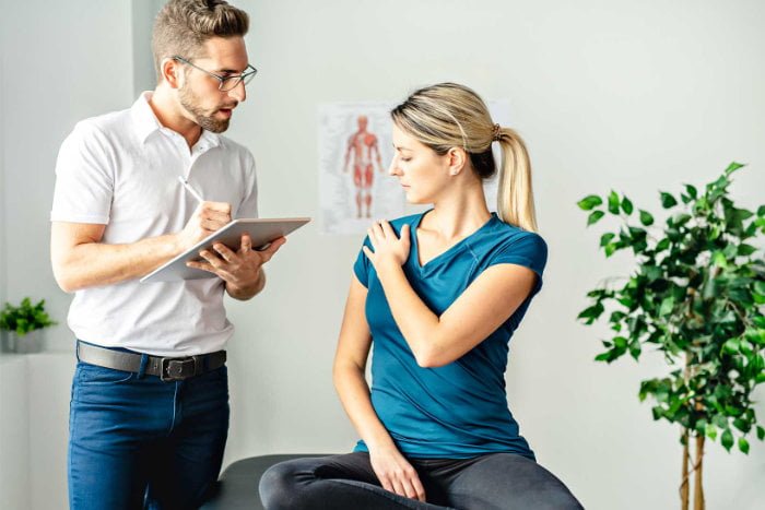 Male chiropractor listening to a female patient pointing to her sore shoulder
