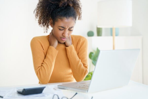 Young woman with chronic pain holding her neck while sitting in front of a laptop