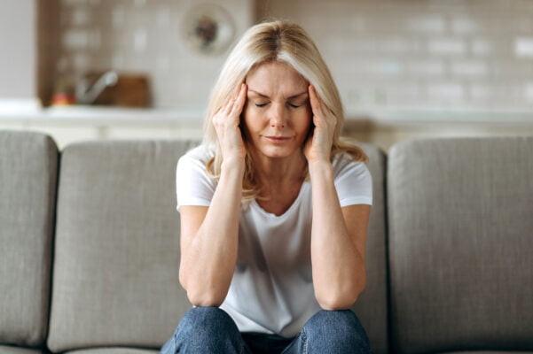 Middle aged blonde woman with a headache sits on couch holding her head with her hands