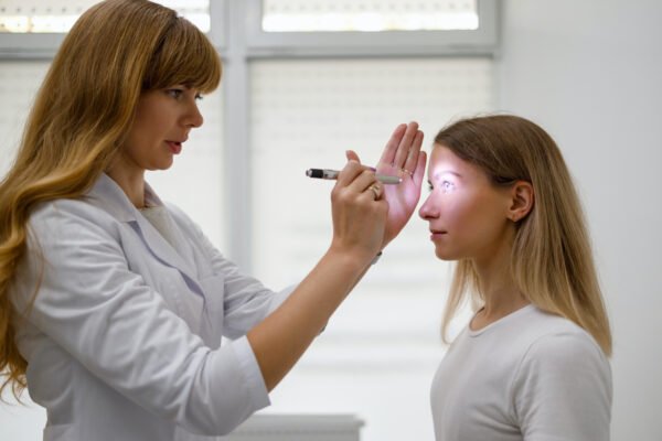 Female doctor checking young female concussion patient's eyes with flashlight.